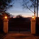 Medium Victorian cast iron pillar light in black, illuminated atop stone gate piers at dusk, showing tapered lantern head with stepped crown and finial.