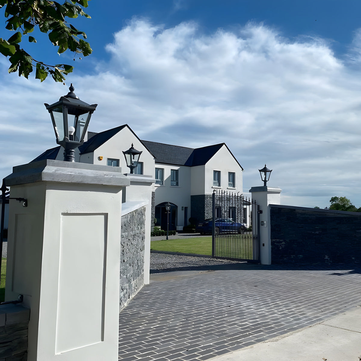 Black Victorian cast iron pillar lights with tapered four-sided heads mounted on flat pier caps at a gated driveway entrance to a white house.