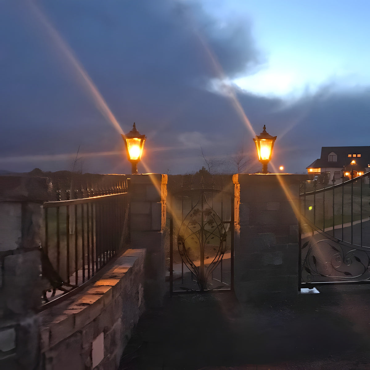 Medium Victorian cast iron pillar lights in black, tapered lantern heads with stepped crowns lit atop matching stone gate piers at dusk.