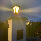 Black cast iron Victorian pillar light with tapered glass head and cast finial, illuminated atop a stone gate post pier cap in a garden setting with railings.
