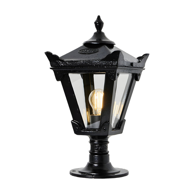 Small Victorian cast iron pillar light in black, tapered four-sided glass head with stepped crown and finial, mounted on pedestal base, lit bulb visible, plain white background.