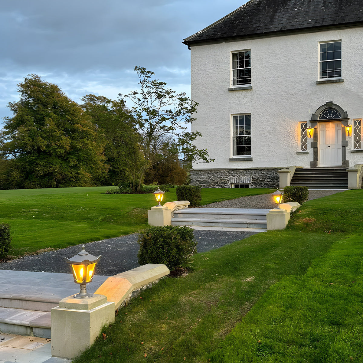 Small Victorian cast iron pillar lights with black finish and lantern-style glass heads, illuminated on stone piers along a garden path leading to a white house entrance.