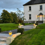 Small Victorian cast iron pillar lights with black finish and lantern-style glass heads, illuminated on stone piers along a garden path leading to a white house entrance.