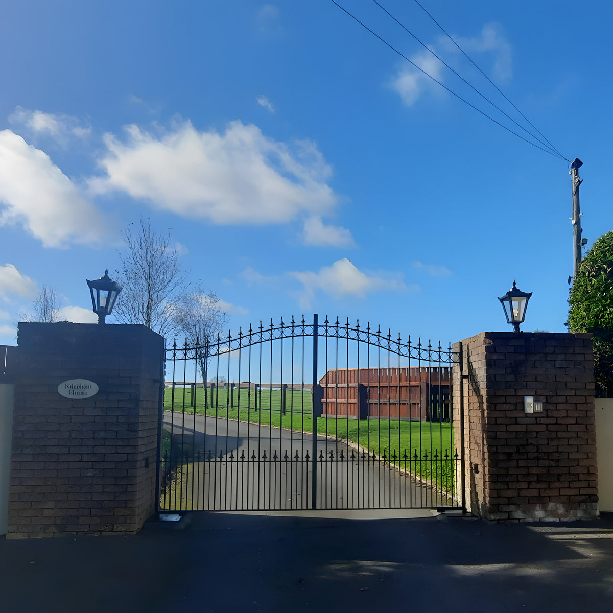 Small Victorian cast iron pillar light in black mounted on a brick gate pier, with a tapered four-sided lantern head and stepped crown atop a stone gateway.