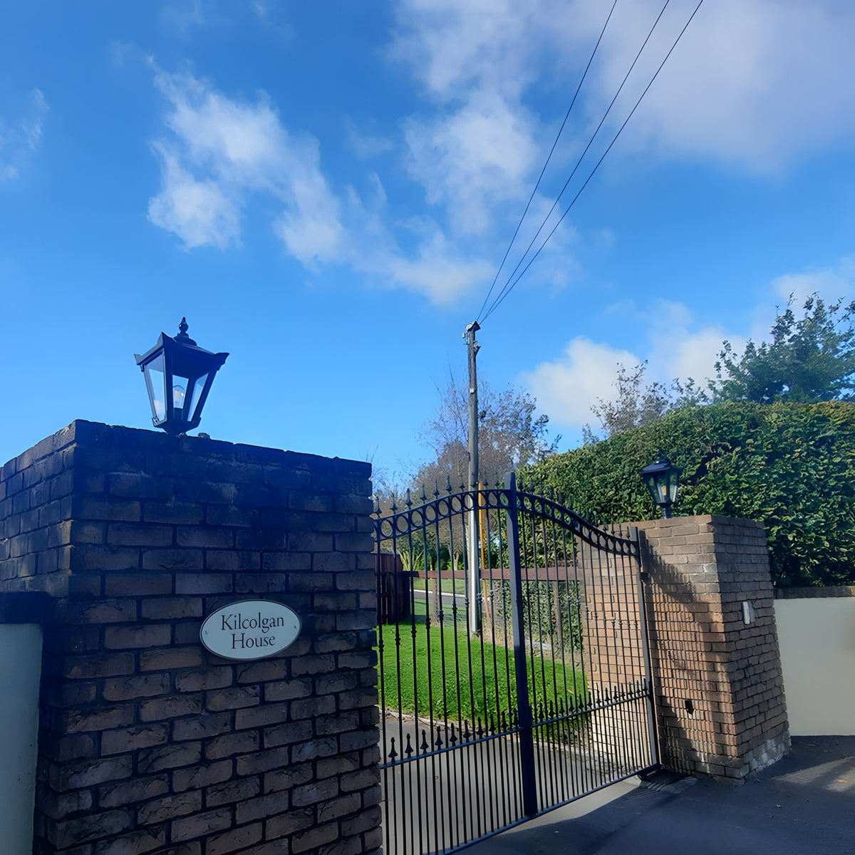 Small Victorian cast iron pillar light in black, mounted atop a brick gate pier with a tapered four-sided head, stepped crown and finial, beside black wrought iron gates.