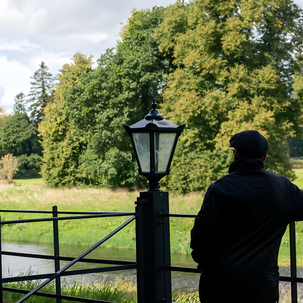 Large Victorian cast iron pillar light in black, tapered four-sided glass head with stepped crown and finial, mounted on a pier post beside a garden fence and riverside setting.