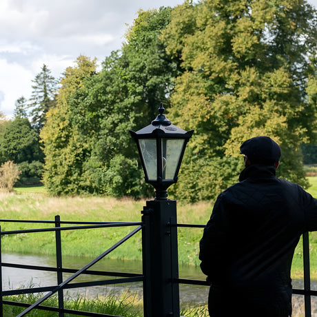 Large Victorian cast iron pillar light in black, tapered four-sided glass head with stepped crown and finial, mounted on a pier post beside a garden fence and riverside setting.