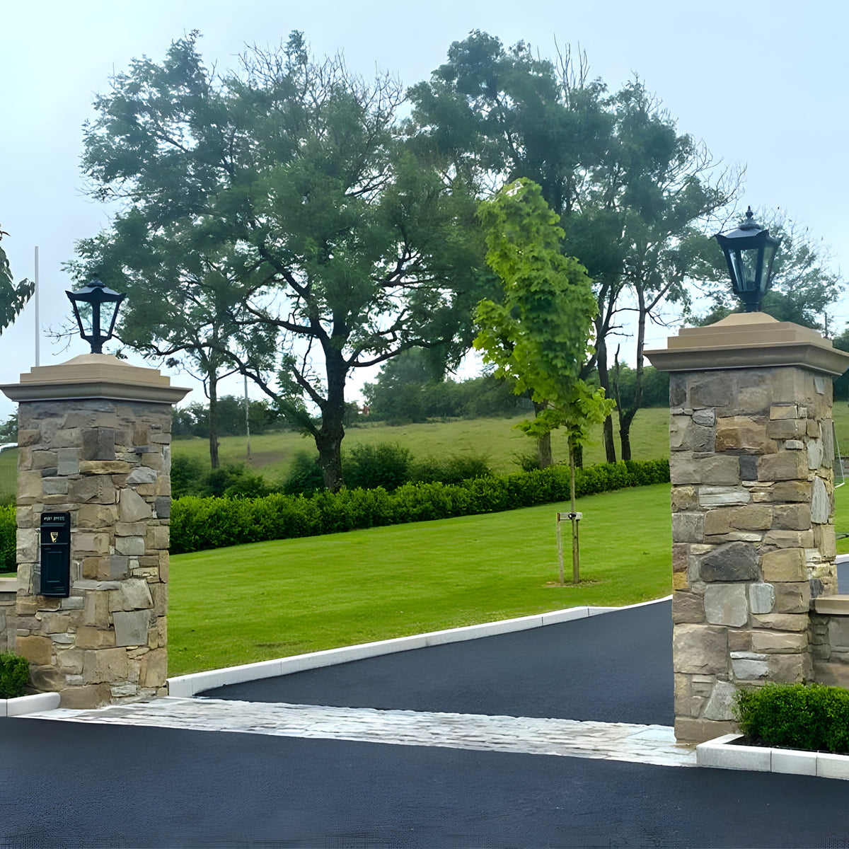 Large Victorian cast iron pillar lights in black atop narrow stone gate piers, tapered four-sided heads with stepped crowns, flanking a driveway entrance.