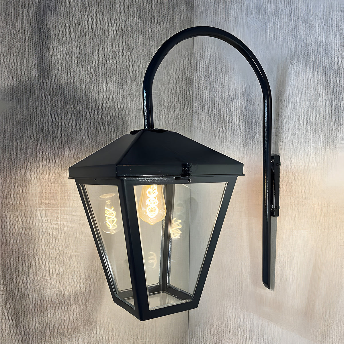 Contemporary downturned wall lantern in black steel with a tapered glass panel shade and curved wall arm, shown illuminated against a plain grey background.