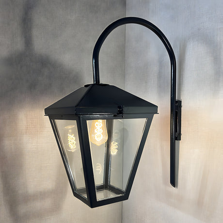Contemporary downturned wall lantern in black steel with a tapered glass panel shade and curved wall arm, shown illuminated against a plain grey background.
