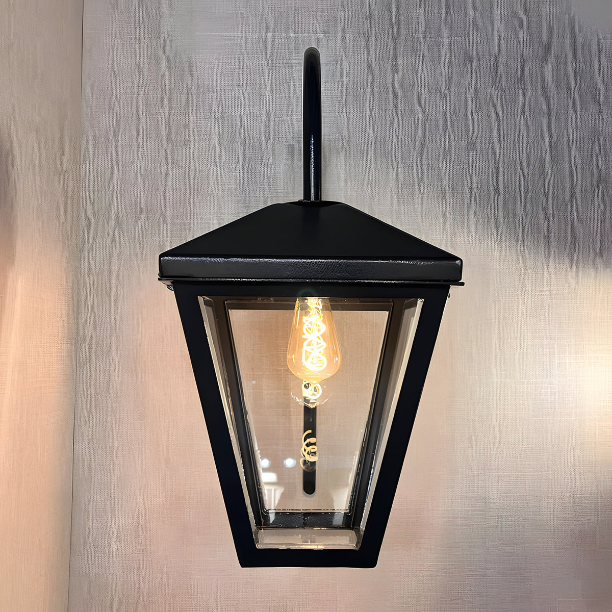 Contemporary downturned wall lantern in black metal with clear glass panels, tapered lantern head, curved wall bracket and exposed E27 bulb, shown lit against a plain light backdrop.