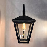 Contemporary downturned wall lantern in black metal with clear glass panels, tapered lantern head, curved wall bracket and exposed E27 bulb, shown lit against a plain light backdrop.