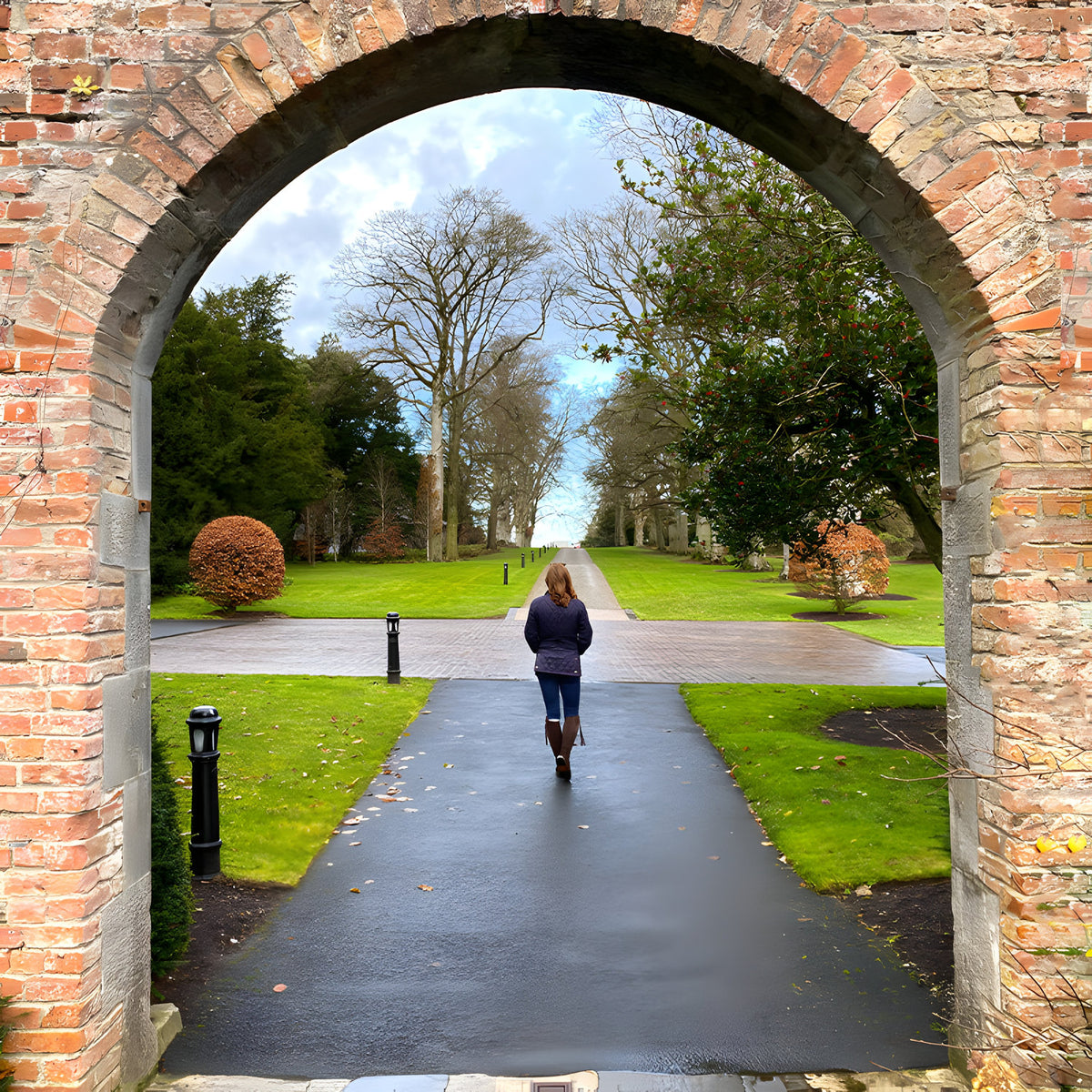 Black cast iron bollard light path marker beside a paved walkway, tall stepped column with frosted lens and domed cap, framed by a brick archway.