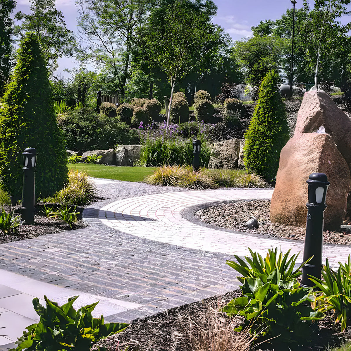 Extra Large Victorian cast iron bollard lights in black lining a garden pathway, with stepped columns and frosted lenses among paved walkways and greenery.