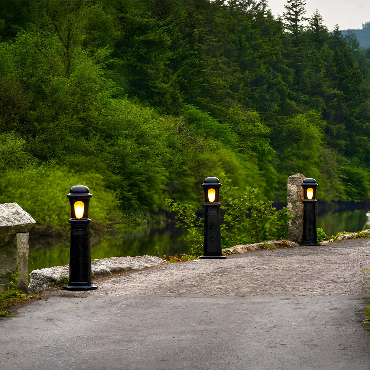 Medium Victorian Cast Iron Bollard Light in black, lit and installed along a stone-edged driveway beside a river, with a traditional stepped column and frosted lens.