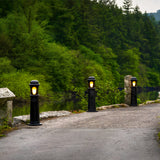Medium Victorian Cast Iron Bollard Light in black, lit and installed along a stone-edged driveway beside a river, with a traditional stepped column and frosted lens.