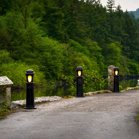 Medium Victorian Cast Iron Bollard Light in black, lit and installed along a stone-edged driveway beside a river, with a traditional stepped column and frosted lens.