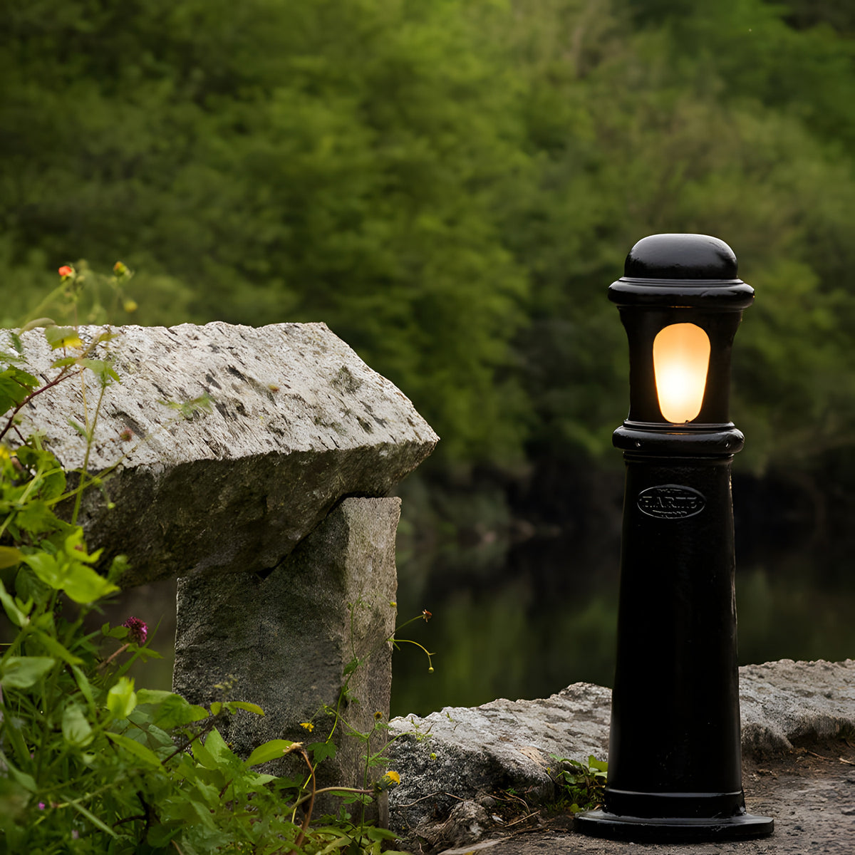 Medium Victorian Cast Iron Bollard Light in black, a stepped column with frosted lens glowing amber, set outdoors beside stone steps and greenery.