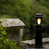 Medium Victorian Cast Iron Bollard Light in black, a stepped column with frosted lens glowing amber, set outdoors beside stone steps and greenery.