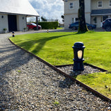 Small Victorian cast iron bollard light in black with frosted lens, illuminated on a lawn edge beside a gravel driveway.