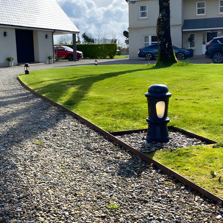 Small Victorian cast iron bollard light in black with frosted lens, illuminated on a lawn edge beside a gravel driveway.