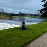 Small Victorian Cast Iron Bollard Light in black with domed cap and frosted lens, installed on a lawn beside a river and footbridge.