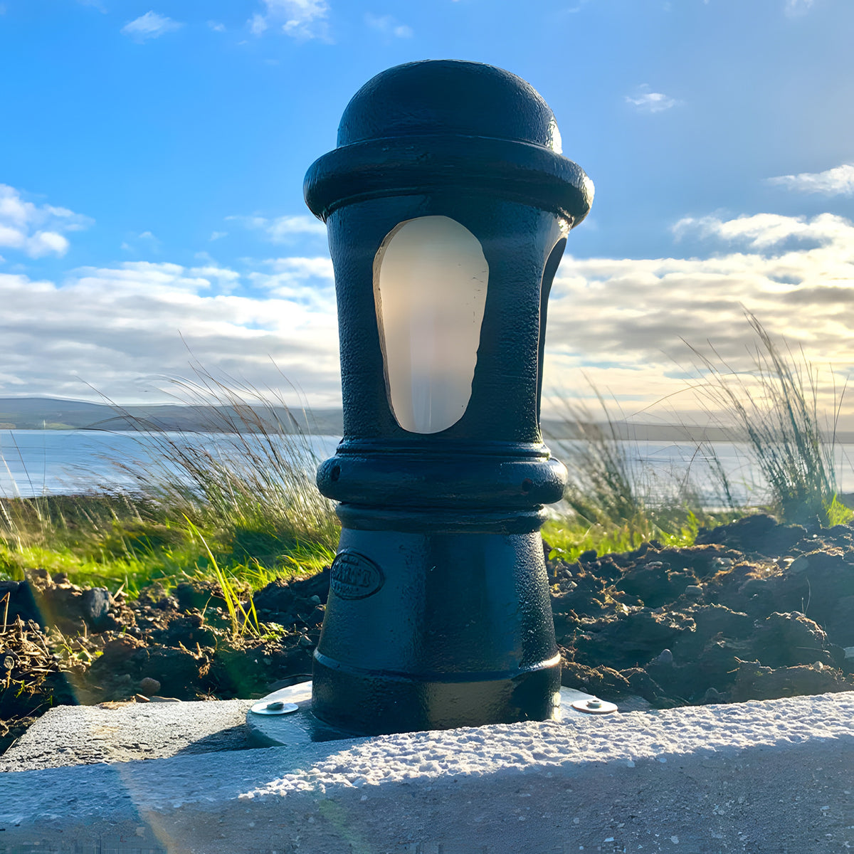 Small Victorian cast iron bollard light in black with a frosted lens, mounted on a stone wall by the coast under a bright sky.