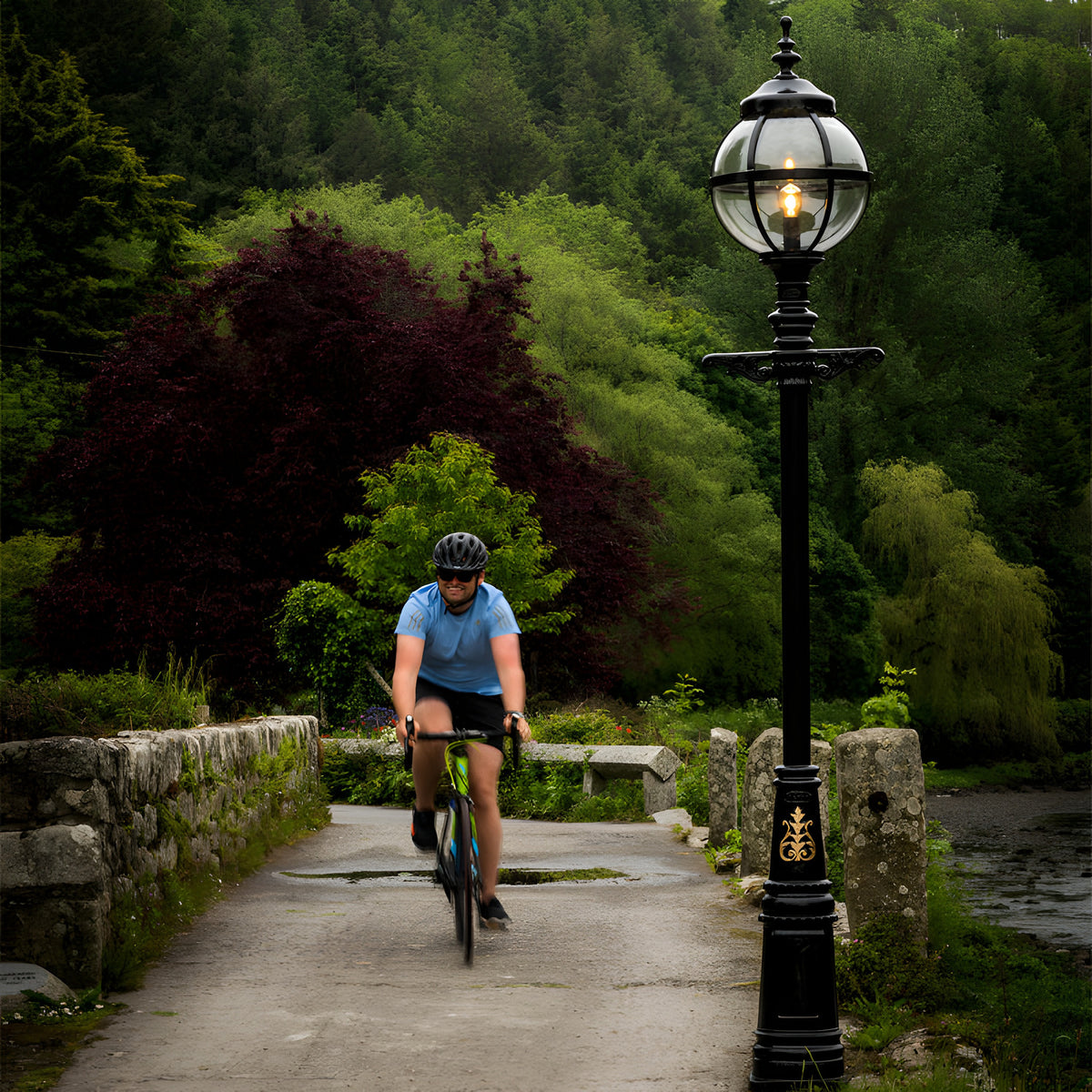 Victorian Cast Iron Globe Lamp Post 3.6m in black, single globe light in a banded cage on a tall ornate pole, shown outdoors beside a stone bridge with a cyclist passing by.