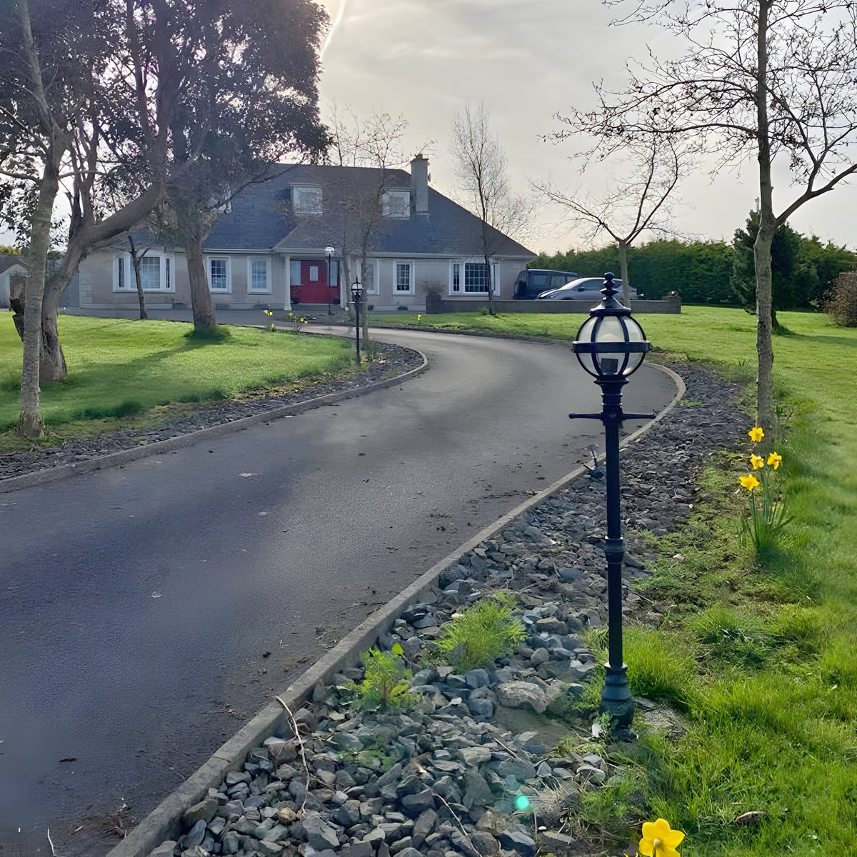 Victorian Cast Iron Globe Lamp Post 1.4m in black, cast iron globe in banded cage on a traditional post, installed beside a driveway edged with gravel and daffodils.