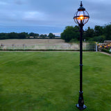 Victorian Cast Iron Globe Lamp Post in black, 2.5m tall with a banded cage globe lit against a dusk garden setting, mounted on a decorative base with a lawn and countryside view behind.