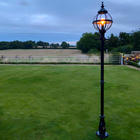 Victorian Cast Iron Globe Lamp Post in black, 2.5m tall with a banded cage globe lit against a dusk garden setting, mounted on a decorative base with a lawn and countryside view behind.