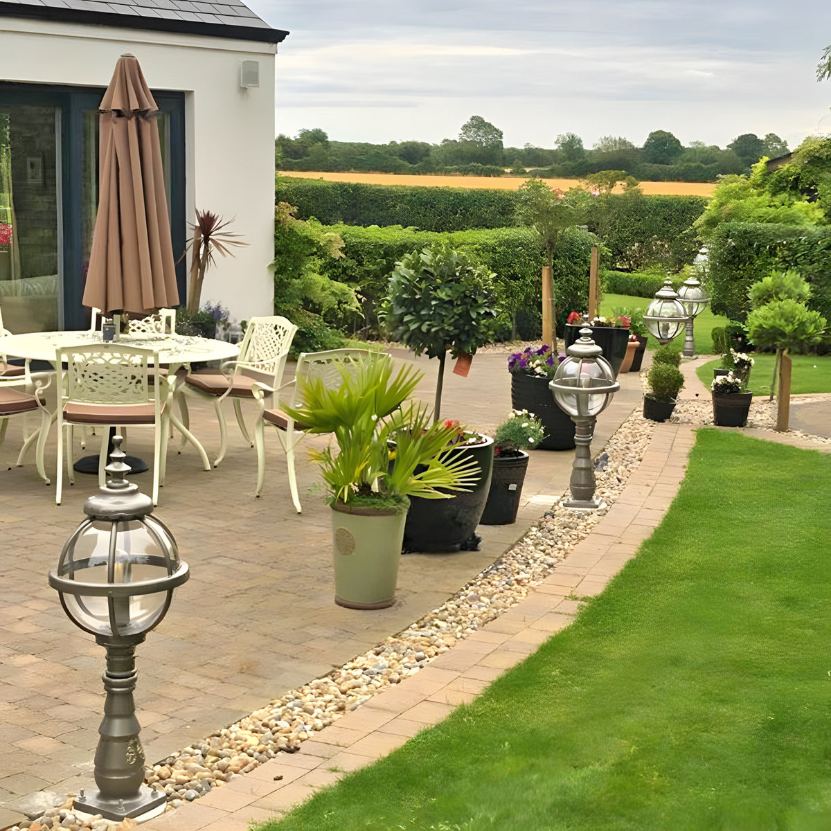 Large Victorian cast iron globe pedestal post light in black, shown lining a garden patio pathway alongside potted plants and outdoor seating.