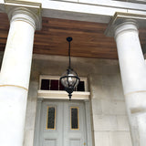 Large Victorian Globe Hanging Lantern in black, cast iron banded cage on a chain, mounted beneath a wooden porch ceiling between two white columns above a grey front door.