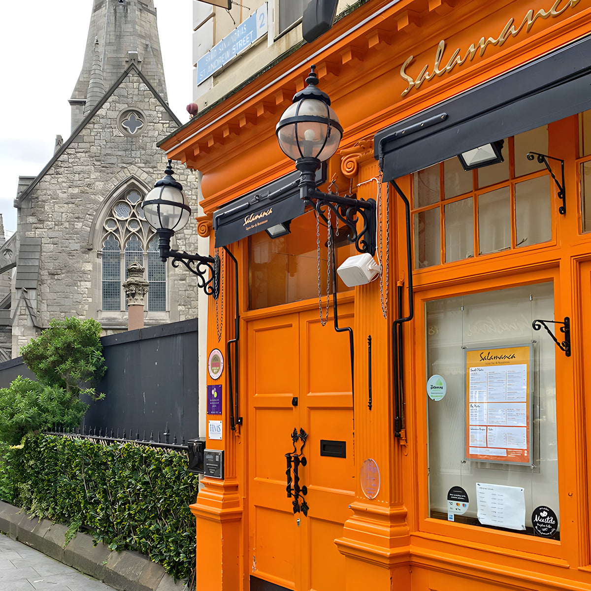 Medium Victorian Globe Wall Lantern in black on a scroll bracket, mounted beside an orange shopfront with a Gothic church visible in the background.