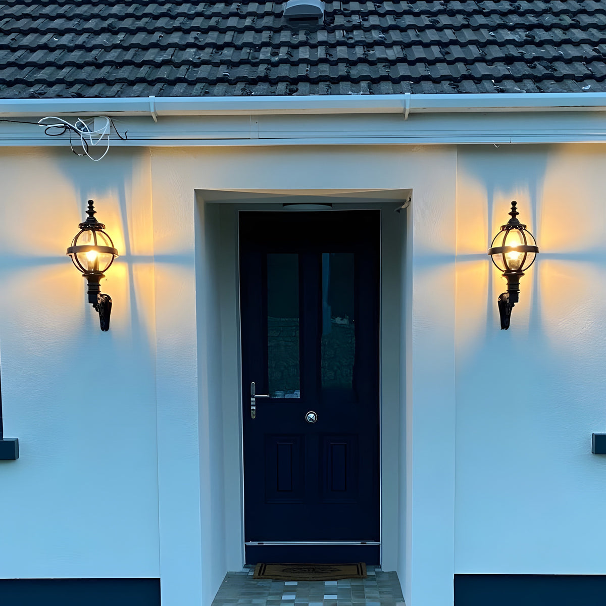 Small Victorian Globe Wall Lantern in black, mounted on a scroll bracket either side of a navy front door, illuminated on a white house exterior.
