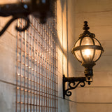 Small Victorian Globe Wall Lantern in black, illuminated on a scroll bracket against wooden panelled wall, showing cast iron cage and glass globe detail.
