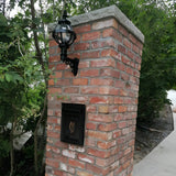 Small Victorian Globe Wall Lantern in black on a scroll bracket, mounted on a brick pillar with a caged globe shade, shown outdoors near greenery.