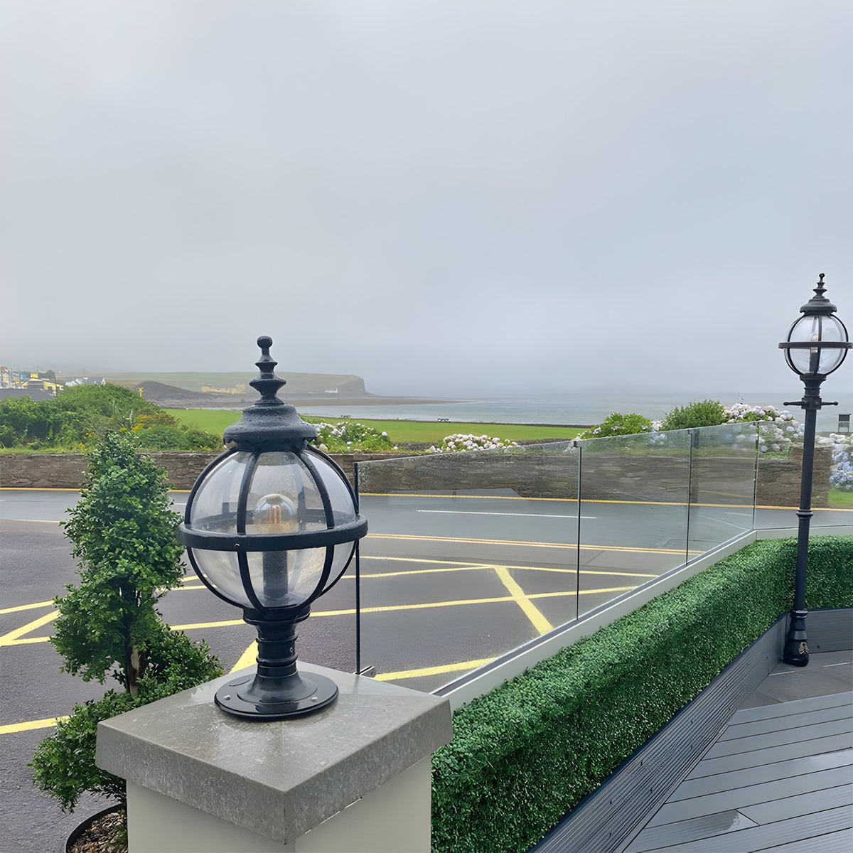Medium Victorian Globe Pillar Light in black, mounted on a stone pier, with a matching lamp post visible along a coastal terrace.