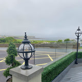 Medium Victorian Globe Pillar Light in black, mounted on a stone pier, with a matching lamp post visible along a coastal terrace.