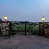 Medium Victorian Globe Pillar Light in black mounted on stone gate piers, illuminated glass globe within a banded cage atop a turned cast base, flanking an ornate iron gate.