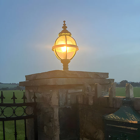 Medium Victorian Globe Pillar Light in black iron, illuminated atop a stone gate pier, showing the banded cage globe against an evening sky.