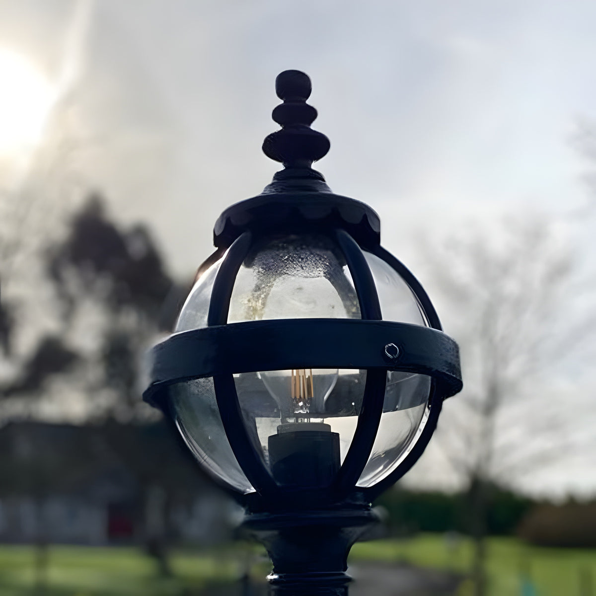 Small Victorian Globe Pillar Light in black, cast iron banded cage with clear globe and visible bulb, ornate finial, shown outdoors against a bright sky.
