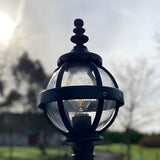 Small Victorian Globe Pillar Light in black, cast iron banded cage with clear globe and visible bulb, ornate finial, shown outdoors against a bright sky.