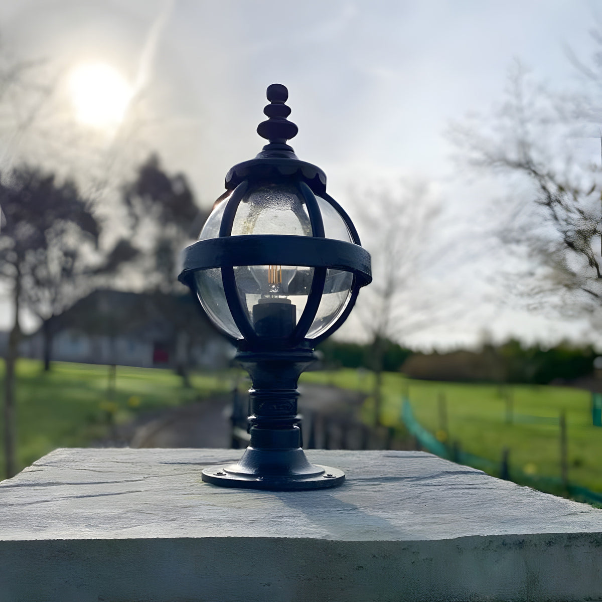 Small Victorian Globe Pillar Light in black, cast iron banded cage with clear globe shade, mounted on a flat pier cap outdoors against a garden backdrop.