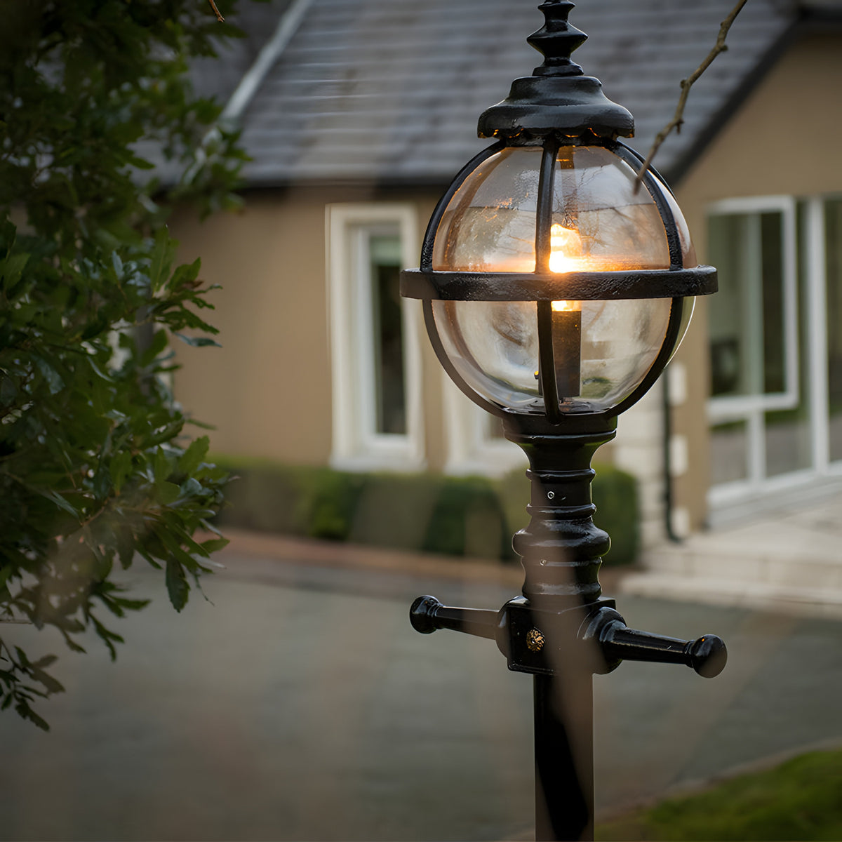 Victorian Manor cast iron globe lamp post in black, banded cage design with lit bulb, shown outdoors on a driveway near a house.
