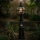 Victorian Manor black cast iron globe pedestal post light with banded cage and polycarbonate glazing, illuminated in a garden setting beside plants and a low wall.