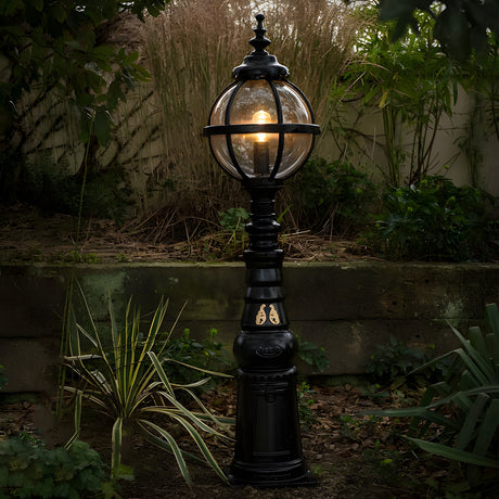 Victorian Manor black cast iron globe pedestal post light with banded cage and polycarbonate glazing, illuminated in a garden setting beside plants and a low wall.