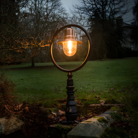 Large railway-style cast iron pedestal post light with copper shade encircled by a round hoop bracket, glowing in a garden setting at dusk.