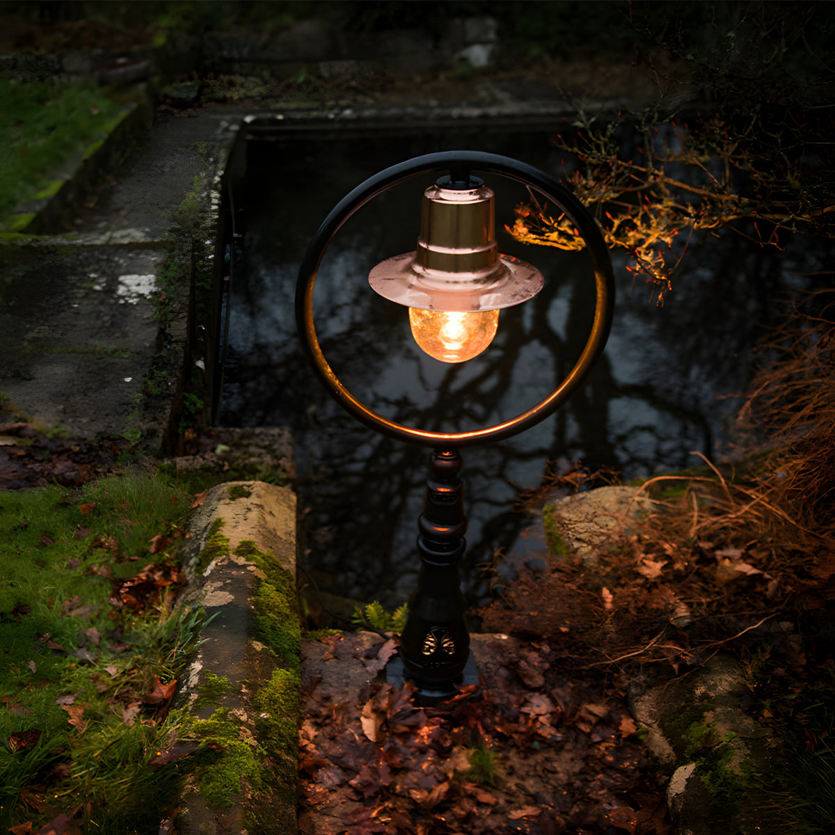 Large Railway Style Cast Iron Pedestal Post Light with copper shade and circular black frame, glowing bulb, set outdoors beside a stone pond amid moss and leaves.