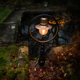 Large Railway Style Cast Iron Pedestal Post Light with copper shade and circular black frame, glowing bulb, set outdoors beside a stone pond amid moss and leaves.